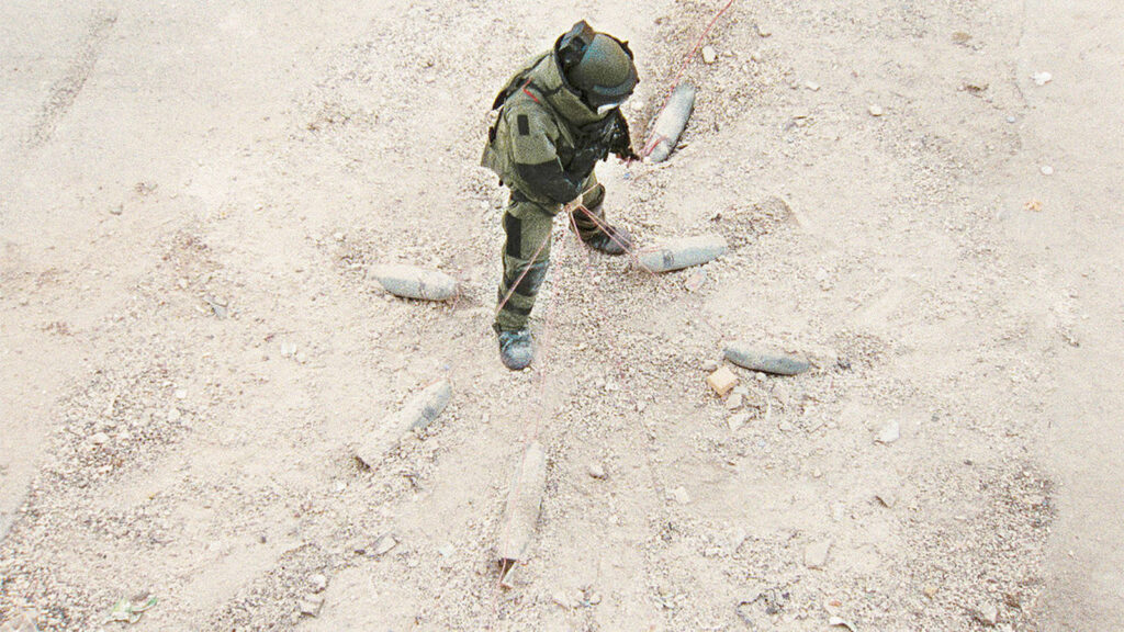 Still from the film "The Hurt Locker". A man in a bomb disposal suit stands surrounded by several bombs wired together.